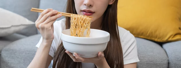 Fotobehang Happy temptation, cute attractive asian young student woman, girl using chopsticks eating instant ramen, noodles soup in bowl in living room at home, cooking meal fast food lifestyle of person. © KMPZZZ