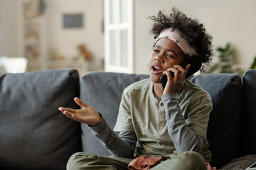 Cute talkative boy with smartphone by ear sitting on sofa in living room and explaining something...