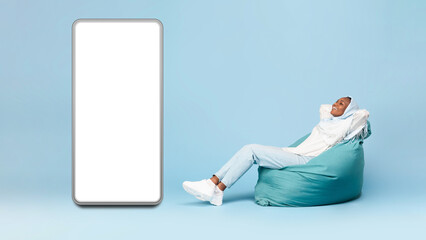 Full length of black muslim lady sitting on beanbag chair next to big blank smartphone with mockup for design © Prostock-studio