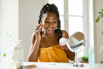 Happy beautiful black woman looking at mirror and applying moisturising face cream, making daily...