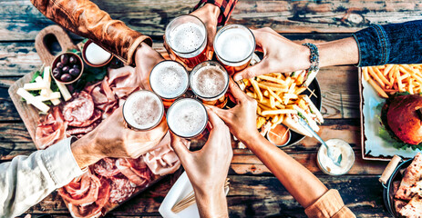 Multi racial friends toasting beer glasses on wooden table covered with food - Top view of people...