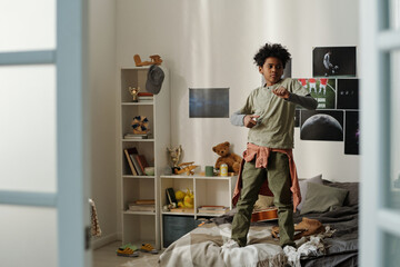 Cute active schoolboy standing on double bed and training defense exercise while practicing karate or some other martial art