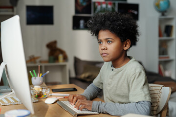 Serious schoolboy sitting in front of computer screen in living room and typing on keyboard while...