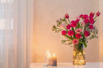 pink flowers in glass modern vase with burning candles in white interior