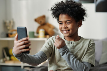 Happy schoolboy holding smartphone in front of himself and taking selfie or communicating in video...