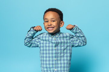 Funny black boy showing biceps and smiling at camera, african american male child demonstrating his power and strength