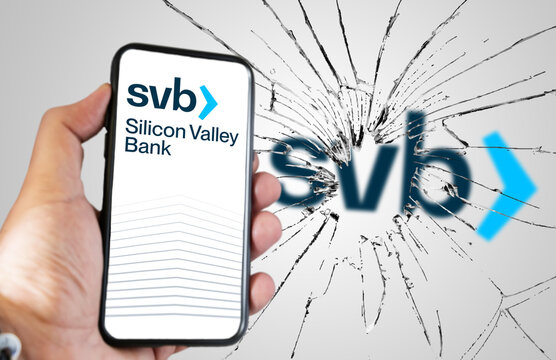 Hand holding a phone with Silicone Valley Bank website on the screen with broken glass and blurred SVB logo in the background