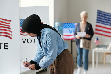 Young Muslim woman in hijab bending over vote booth and making her choice of candidate during presidential elections at polling place - obrazy, fototapety, plakaty