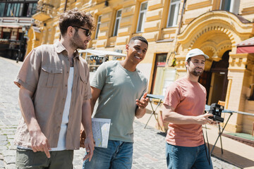 joyful interracial travelers with city map and vintage camera walking with tour guide in sunglasses on Andrews descent in Kyiv.
