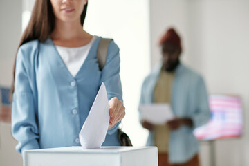 Focus on hand of young female voter putting ballot paper into box while standing in front of camera against black man in polling place