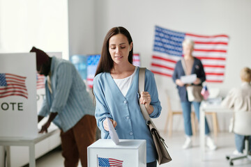 Young brunette woman in casualwear putting ballot paper into box with American flag while voting...