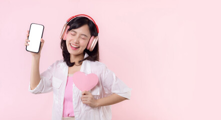 Happy cheerful smiling asian woman with hand holding heart and earphones showing blank screen mobile phone or new smartphone music application advertisement mockup isolated on pink studio background.