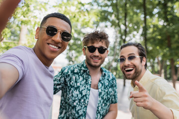 Positive interracial friends in sunglasses pointing at camera in summer park.