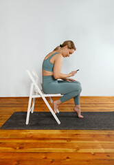 A young blonde girl in green sportswear is sitting on a white chair using her phone after doing yoga exercise. The concept of sports and correct posture for girls.