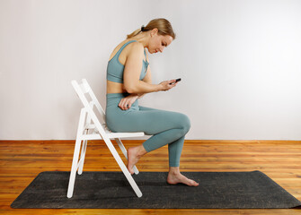 A young blonde girl in green sportswear is sitting on a white chair using her phone after doing yoga exercise. The concept of sports and correct posture for girls.