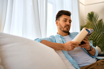 Mid adult man reading book while relaxing on sofa at home.