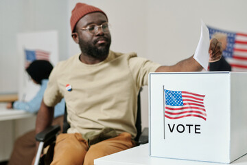 Box for ballot papers on desk and young African American man with disability sitting in wheelchair...