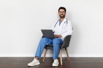 Male Therapist Using Laptop While Sitting In Chair Over White Wall Background