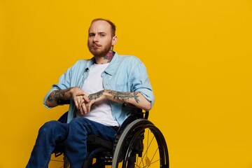 A man in a wheelchair problems with the musculoskeletal system looks at the camera thoughtfully with tattoos on his arms sits on a yellow studio background, health concept a person with disabilities