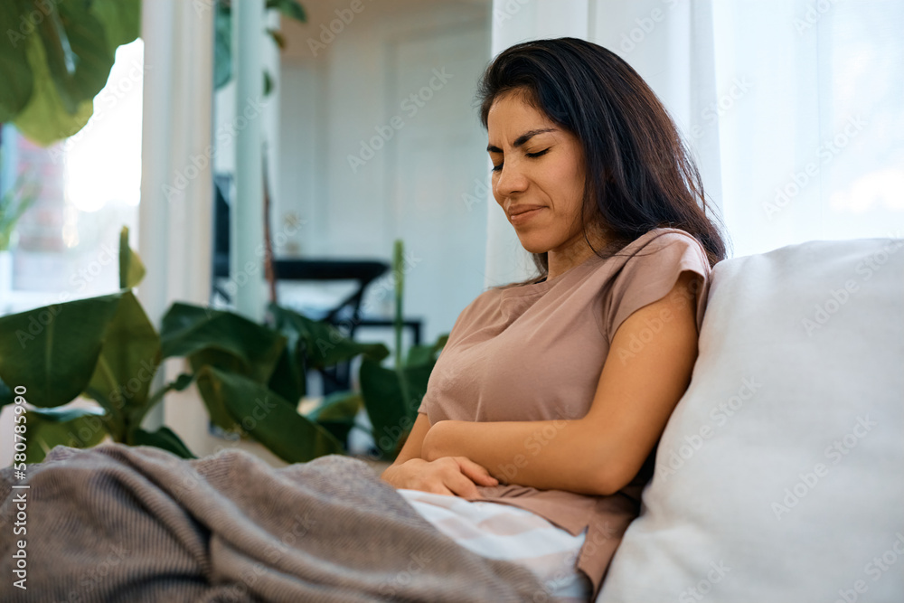 Wall mural woman suffering from stomach pain while sitting on sofa in living room.