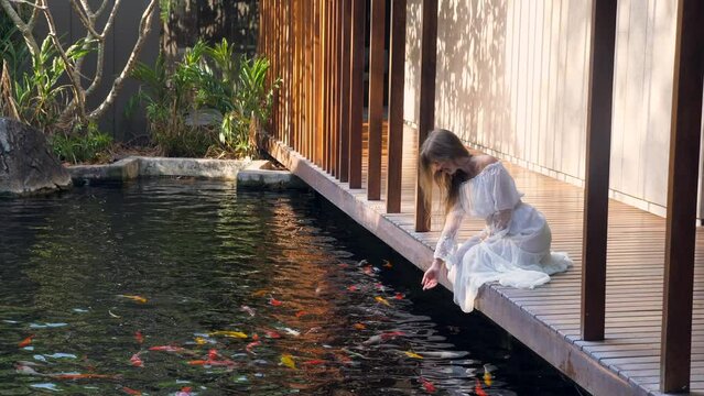 Slow motion footage of woman in white dress relaxing at luxury spa with fish pond. Sitting on wooden bridge and enjoying nature. Zen, meditation.