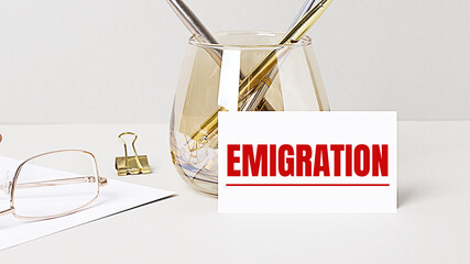 Work desk with gold glasses, pen, white card with the text EMIGRATION. Business concept. Home Office. Workplace close-up.
