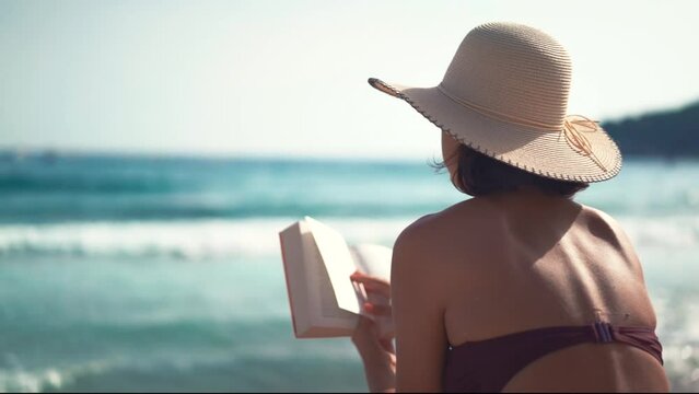 Beautiful young female with hat reading book on sandy beach enjoying her holiday next to sea. What book to read on vacation? Reading books on beach.