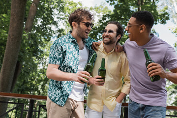 carefree multicultural friends in stylish summer outfit embracing while holding beer in city park.