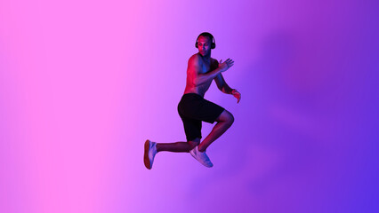 Fototapeta na wymiar Athletic African American Male Jumping Wearing Headphones Over Purple Background