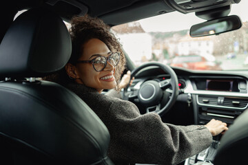 Woman driving car with one hand on steering wheel and another on gear lever and looking at camera