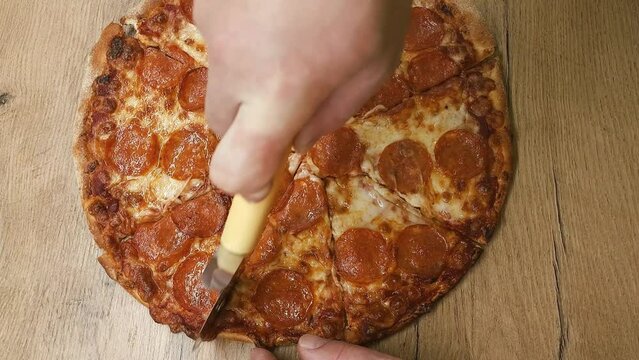Top view of a man hand cutting pizza with a round cutter knife on the wooden table in the kitchen