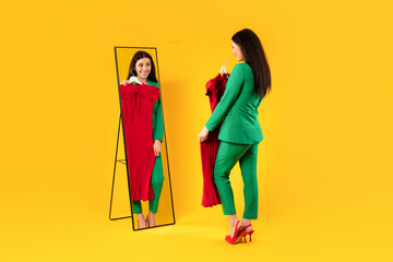 Happy european woman holding red dress and looking at her reflection in mirror, trying on new...