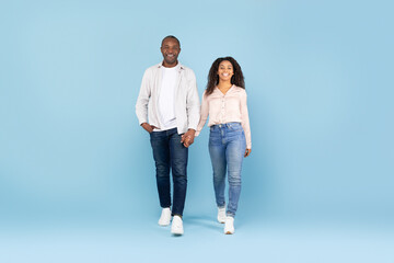 Fototapeta premium African american couple holding hands and walking towards camera, blue studio background, full body length, copy space