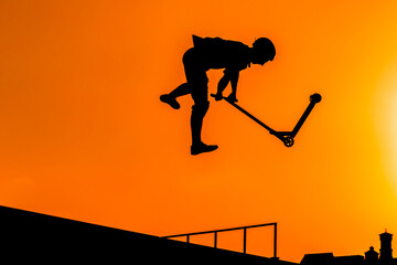Unrecognizable teenage boy silhouette showing high jump tricks on scooter against orange sunsetwarm sky at skatepark. Sport, extreme, freestyle, outdoor activity concept