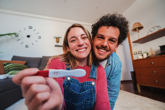 Happy caucasian young couple smiling excited showing at camera their positive pregnancy test at home. Husband and wife celebrating that they are expecting a baby holding a predictor in their hands