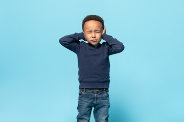Unhappy black little kid boy closing eyes and ears being annoyed with noise, standing isolated over...