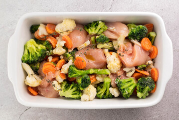 Raw chicken breast, broccoli, cauliflower, carrots, thyme sprigs on a gray table in a ceramic baking dish. The concept of cooking healthy food.