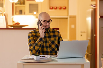 Freelance and online communication. Caucasian bald bearded young man talks on cellphone and works on laptop. Concept of remote work and freelance