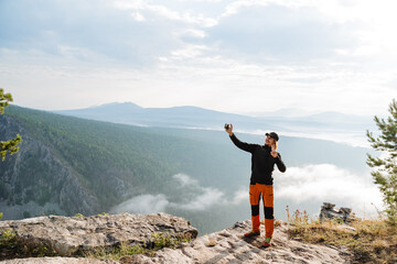 Naklejka na ściany i meble A guy takes a selfie in the mountains against the background of clouds, a mountain trip alone, a tourist shoots himself on a smartphone camera, a man on a hike.