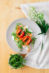 Vertical top view of rocket salad with tomatoes, carrots and yogurt sauce on a grey plate, with flowers and kitchen towel over a wooden table
