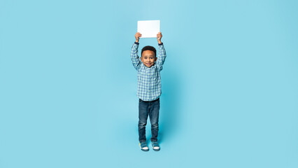Full length shot of african american little boy holding blank card and smiling to camera, posing over blue background