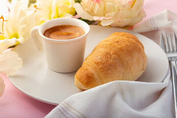 Croissant on a white plate with a cup of espresso coffee with a heart on it, flowers and pink background