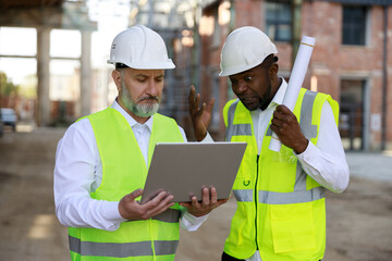 Fototapeta premium Caucasian senior foreman with wireless laptop discussing, showing about all peculiarities of project with african american male architect at construction area.