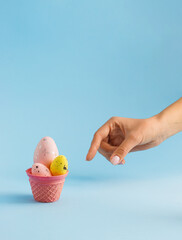 Woman's hand and ice cream cone with colorful eggs on pastel blue background. Minimal Easter concept. Happy Easter card.
