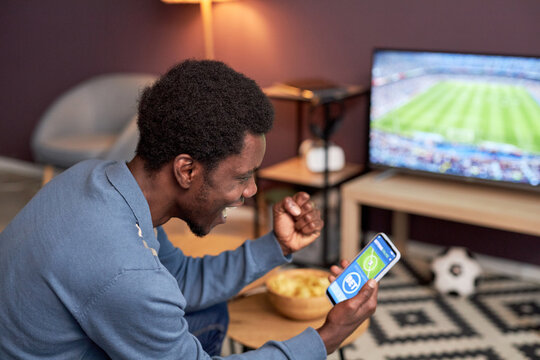 Side view portrait of black man holding smartphone with online sport bets app and cheering for football team
