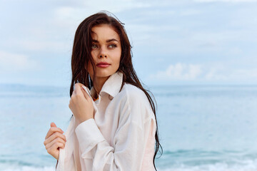 Fototapeta na wymiar Portrait of a beautiful pensive woman with tanned skin in a white beach shirt with wet hair after swimming on the ocean beach sunset light with clouds, the concept of freedom and mental health