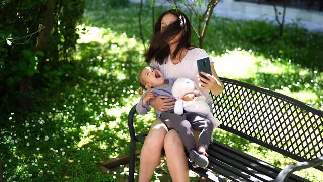 Mom with a little girl take a selfie on a smartphone while sitting on a bench in the garden