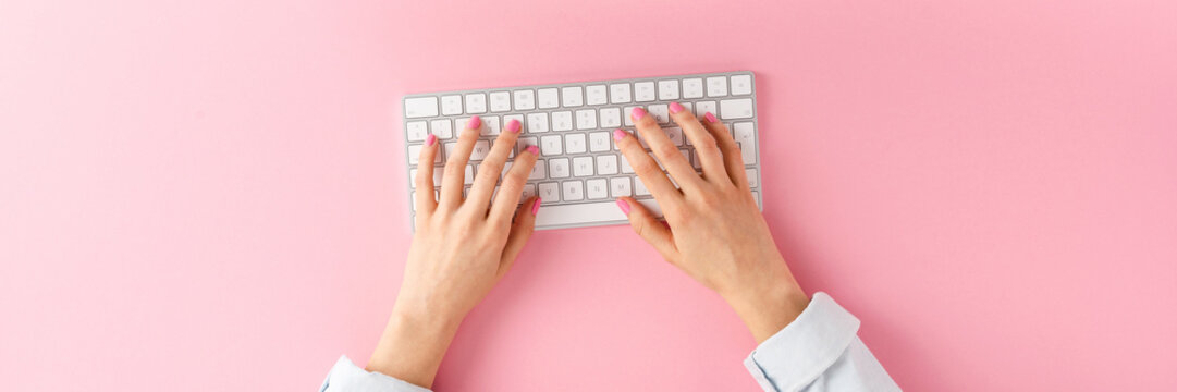 Office desktop concept. Woman&rsquo;s hands working on computer on pink background. Top view