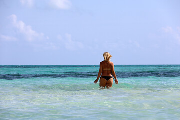 Tanned woman with blond hair wearing bikini going to swim in azure sea water, back view. Beach vacation on Caribbean islands