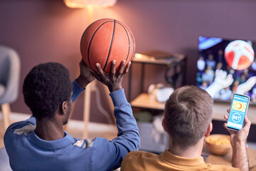 Back view at young black man holding basketball ball while watching sports match at home with friends